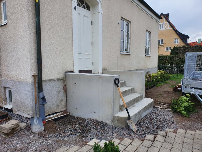 Oformade betongtrappor vid husvägg med skottkärra, spadar och nyligen borttagna växter i trädgården.