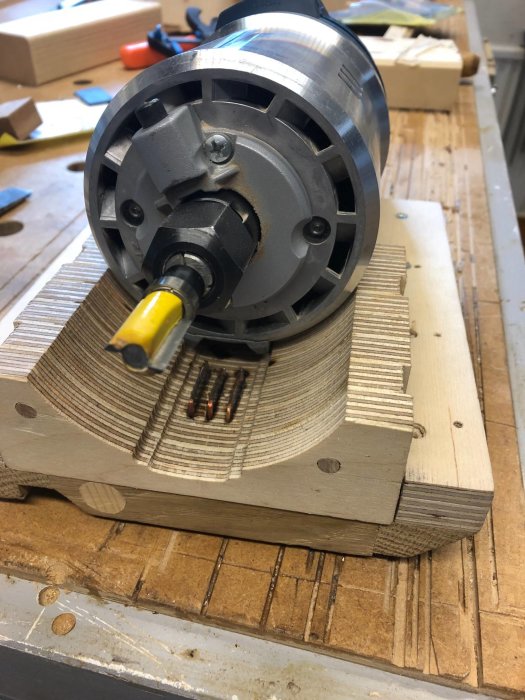 Fräsmotor fastspänd i en nyligen fräst träadapter på en arbetsbänk med verktyg i bakgrunden.