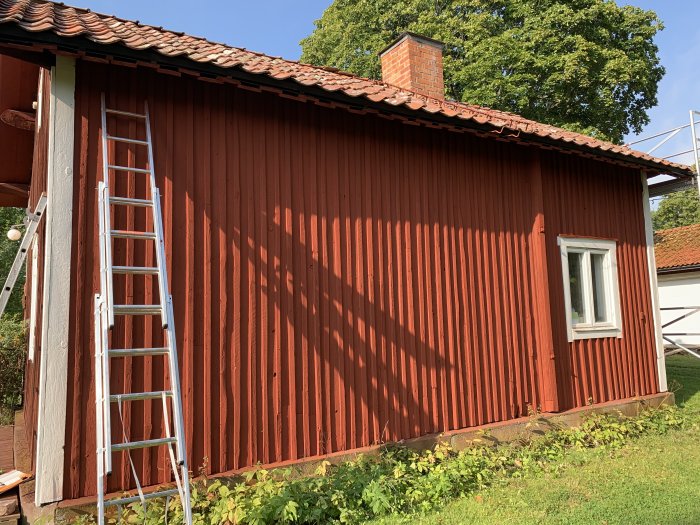 Rödmålat trähus med stege lutad mot väggen och skuggor från tidig morgonsol.