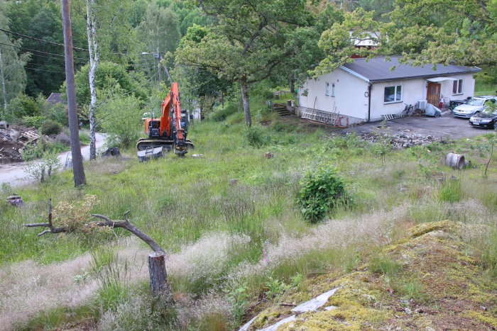 Grävmaskin på en rensad tomt med en vit enplansvilla och bilar i bakgrunden, redo för byggstart.