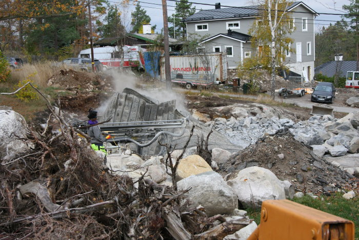 Person använder sprängutrustning för att ta bort berg på en byggplats med hus och fordon i bakgrunden.