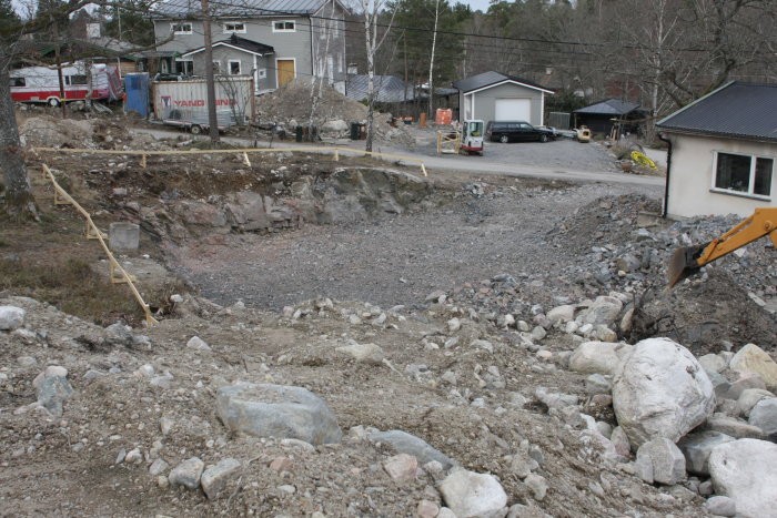 Schaktbotten av jämnt grus med grävmaskinsarm i kanten, omgivet av hus och byggmaterial.