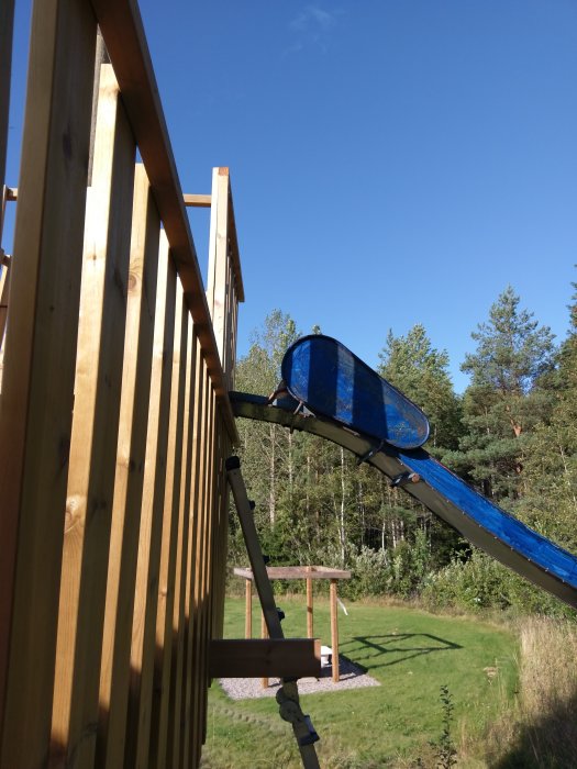 En blå rutschkana utan sidor i början fäst vid en träkonstruktion utomhus.