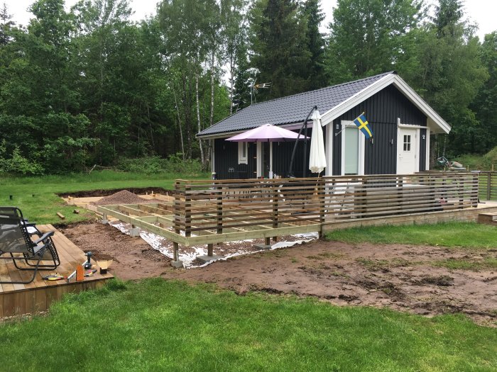 Svart trästuga som blivit permanentboende med nybyggd terrass och svensk flagga.