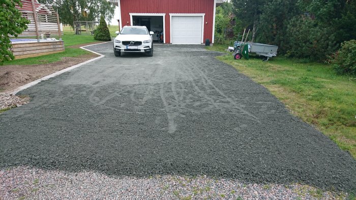 Bil parkerad på nygrusad uppfart framför röd garagebyggnad med öppen port och grönska runtomkring.