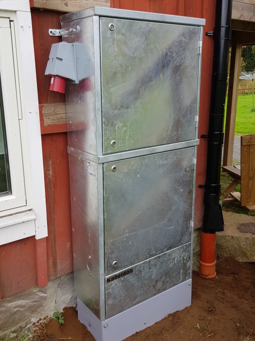 Metallskåp för elinstallation märkt med "Kabeldon", placerat utomhus mot en röd trävägg, med skyddsrör och jordning.