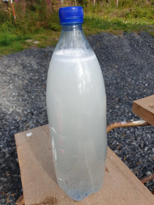 Plastflaska med mjölkigt vatten från nyligen borrade brunn, stående på träplank utomhus.