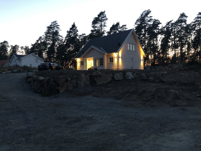 Nytt vitt hus med utomhusbelysning i skymningsljus, omgivet av skog och stenig mark.