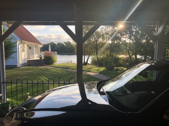 Utsikt från carport mot sjö med hus och soluppgång reflekterad på bilens tak.