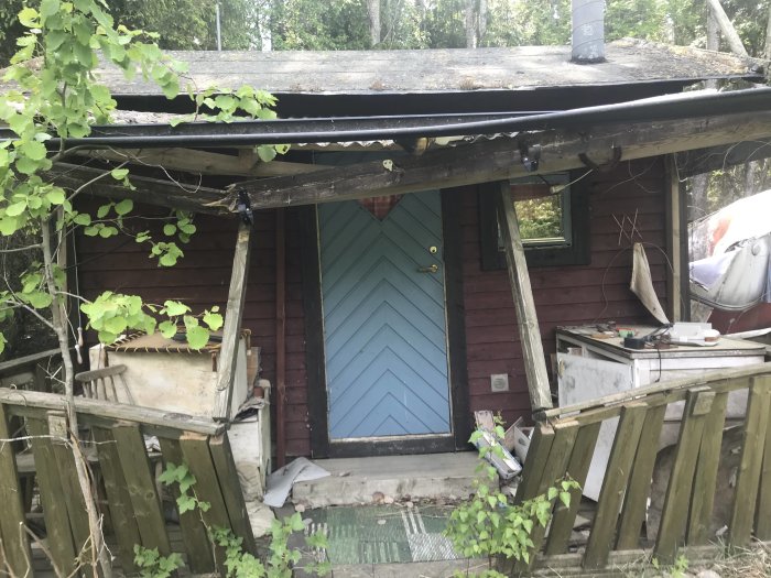 Förfallen stuga med igenväxt ingång och skräp runtomkring, inklusive en gammal husvagn.