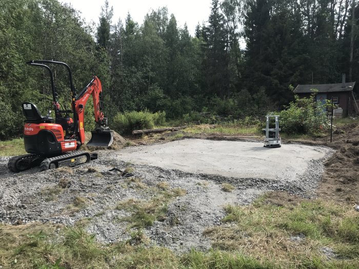 En nyligen grävd och grusbelagd yta för byggprojekt med en minigrävare till vänster och en skogsbakgrund.