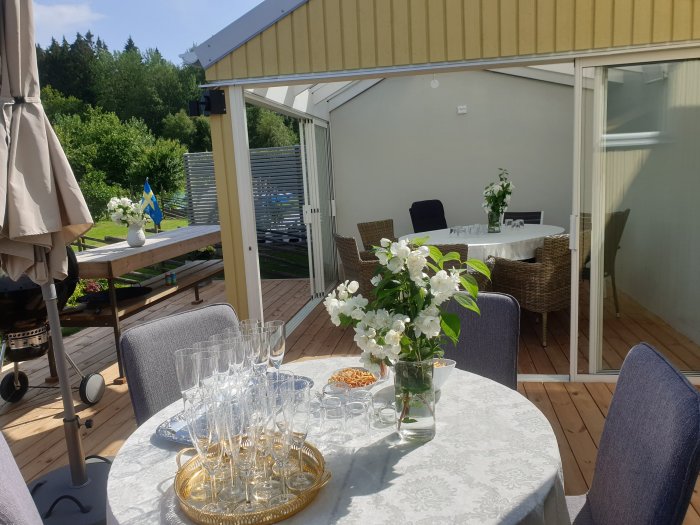 Soligt uterum med dukat bord, glas och blomvas framför terrassmöbler och öppen glasdörr.