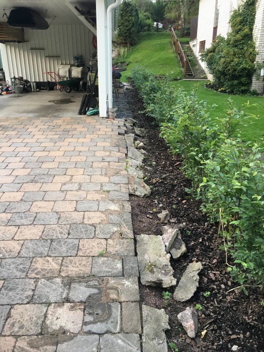 Marksten-uppfart med skadade, lösa kanter och synlig geotextil vid hus med carport och trädgård.