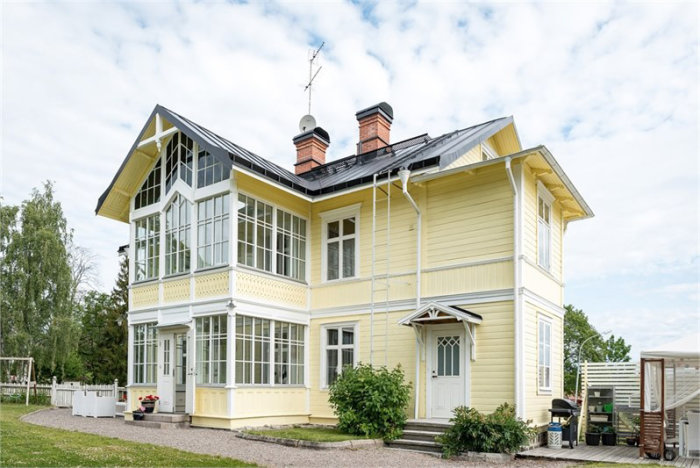 Gult äldre trähus med veranda och stora fönster omgivet av grönska.