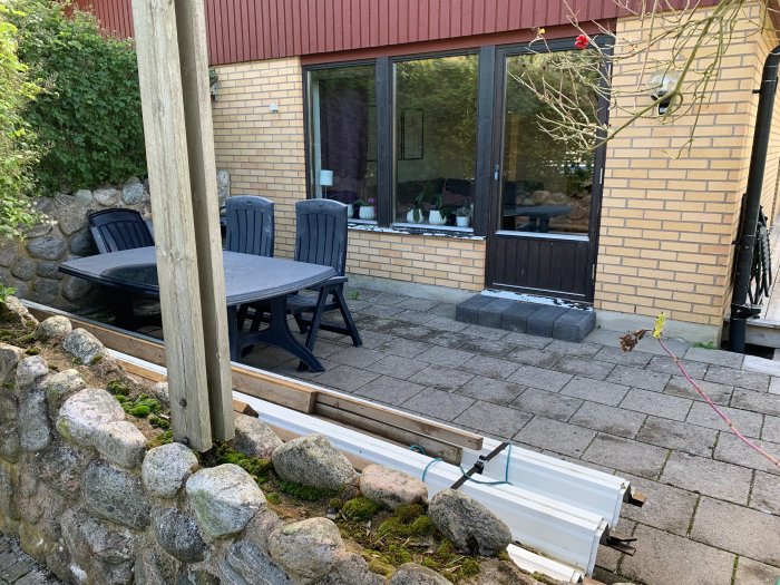 Stenlagd terrass med gammal stenmur, svarta utemöbler och hus med fönster samt byggmaterial i förgrunden.