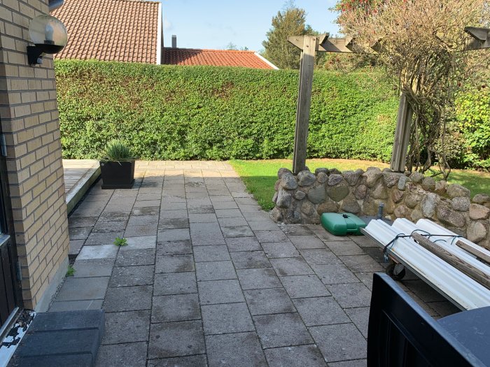 Stenlagd terrass med en mur i behov av renovering, angränsande till en befintlig altan och en grön häck.