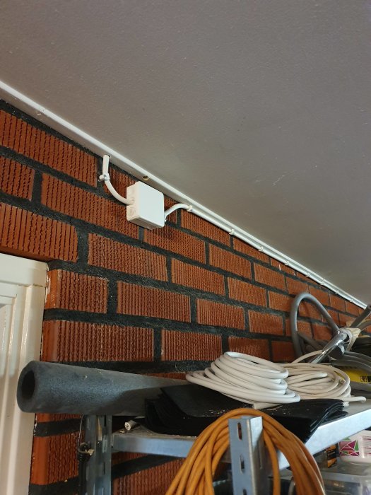 Elinstallation med kapsling och ledningar monterade högt upp på en tegelvägg ovanför en hylla med diverse byggmaterial.