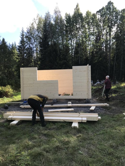 Två personer bygger en träkoja i skogen, stommen är uppe och timmer ligger på marken.