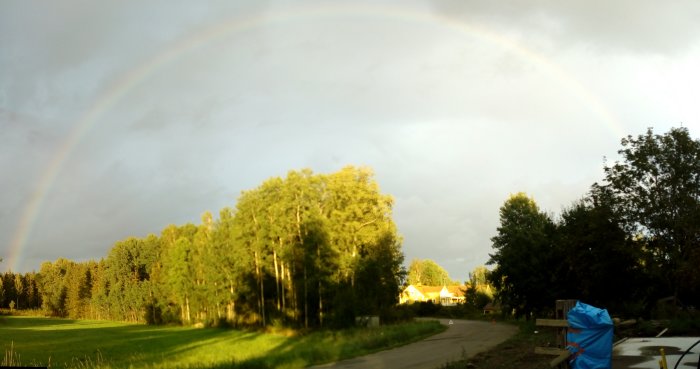 Fullständig regnbåge över ett landskap med gröna träd och ett hus på en solig eftermiddag.