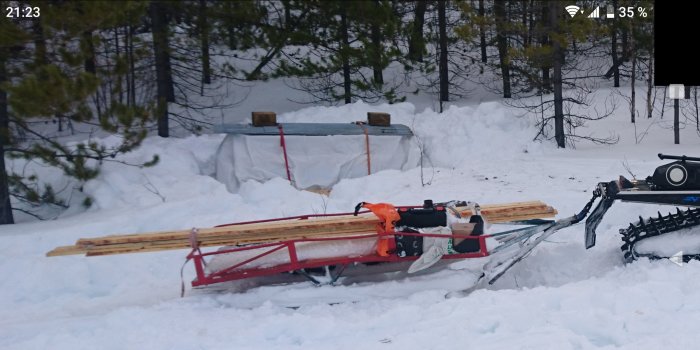 Byggmaterial och verktyg på en pulka i snötäckt skogsområde, nära en tillfällig jaktkoja.
