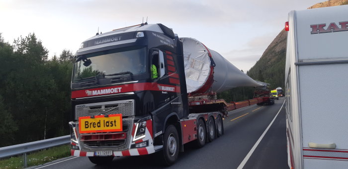 Specialtransport av en stor vindturbinvinge på en lastbil på en väg omgiven av skog och berg i Norge.