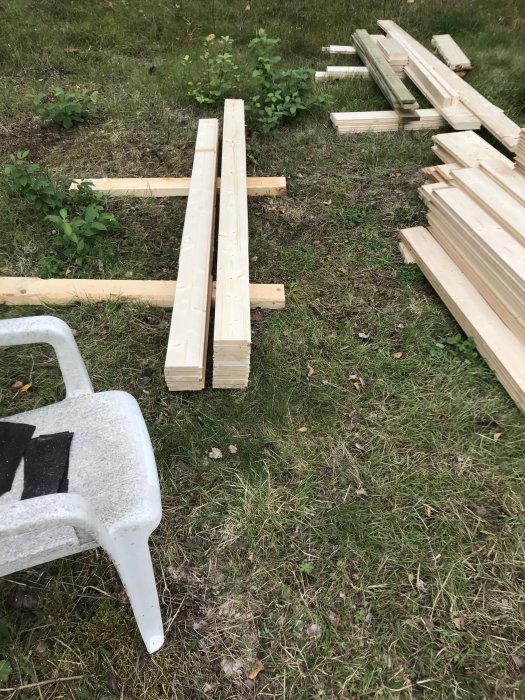 Träbrädor av olika storlekar och en plaststol ordnade på gräs för ett byggprojekt.