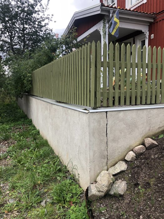 L-formad stödmur med 3 cm glipa där en del ansluter till huset, med staket ovanpå och svensk flagga.