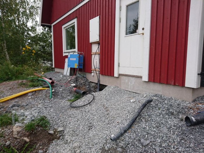 Provisoriskt blått byggströmskåp vid röd husvägg med ledningar och grusyta.