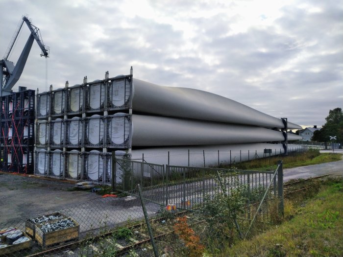 Vindturbinblad som transporteras på lastningsområde med stora kranar i bakgrunden.