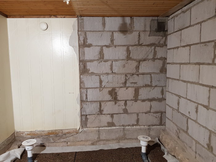 Hörn av ett rått badrum under renovering med synliga VVS-installationer och delvis putsade betongväggar.
