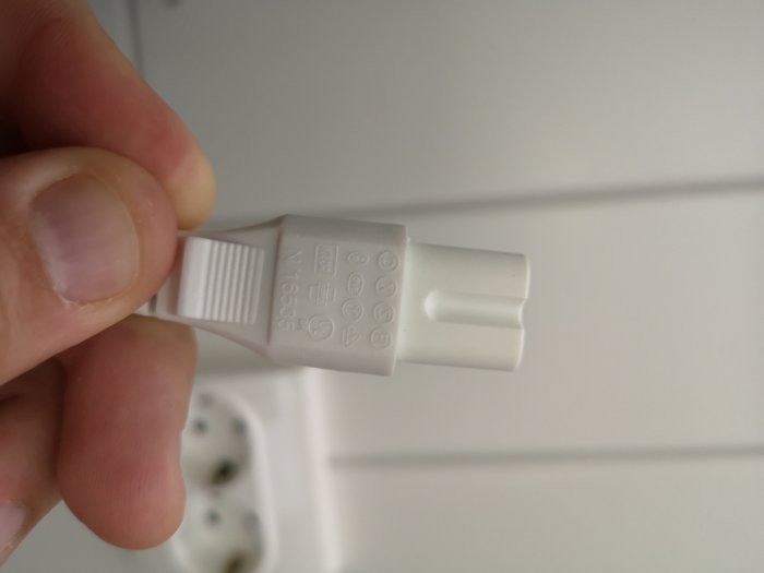 Hand håller en vit 230v elkontakt med sladd framför en vit vägg och ett oanvänt vägguttag.