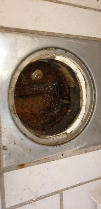 Smutsig golvbrunn omgiven av kakel, med synliga avlagringar och missfärgningar, indikerar underhållsbehov.