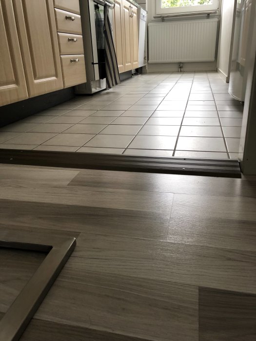 Köksgolv med grått klinker intill en övergång till laminatgolv, under skåpstommar och vid en vit radiator.