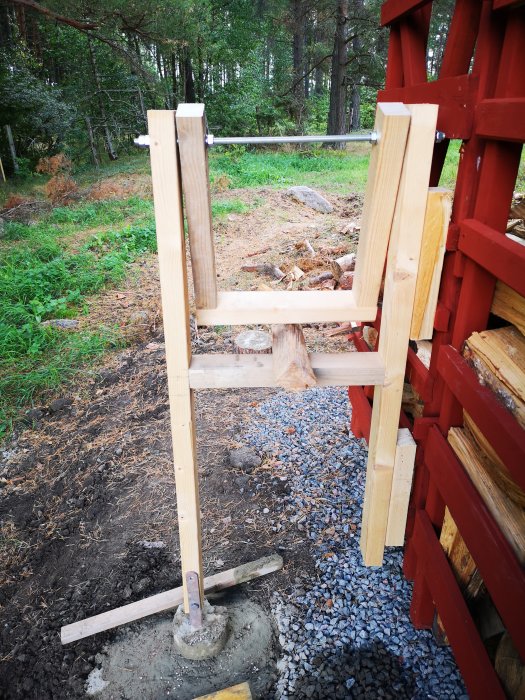 Hemmagjord sågbock av återbrukade trädelar framför en rödmålad byggnad och grönskande skog.