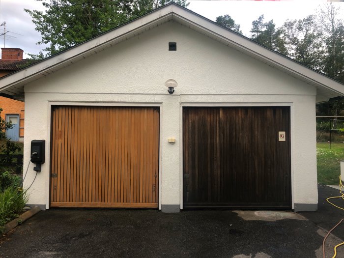 Dubbelgarage med en nymålad vit fasad och två garageportar, en oljad i teaknyans och den andra mörk och obehandlad.