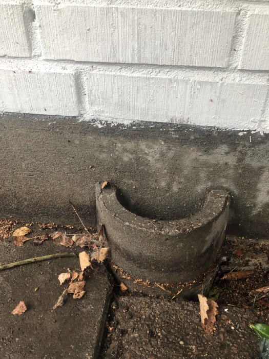 Betongventil nära grunden på ett hus, med tecken på läckage och nedfallna löv runtomkring.
