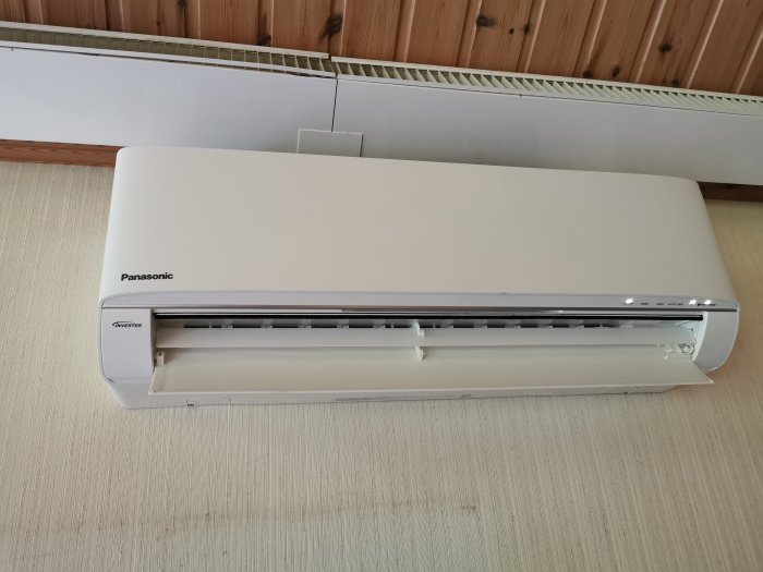 Nyinstallerad Panasonic HZ25UKE luftvärmepump monterad på en vardagsrumsvägg, ovanför en radiator och under ett träpaneltak.