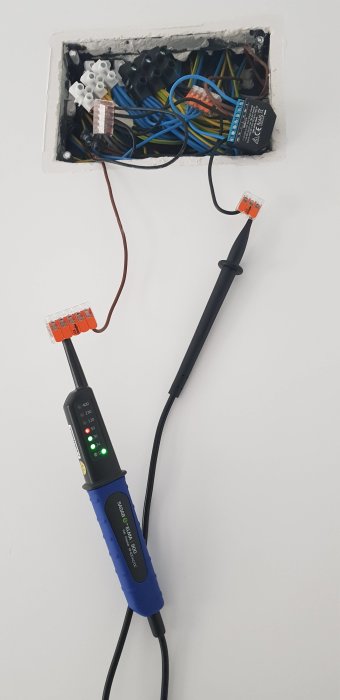 Öppen eldosa med kablar och kopplingsklämmor, och en voltmätare som visar mätvärden ansluten till en kabel.