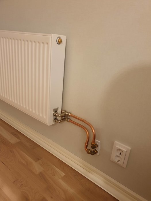 Vit radiator uppsatt på vägg med exponerade kopparledningar ovanför golvlisten och intill eluttag.