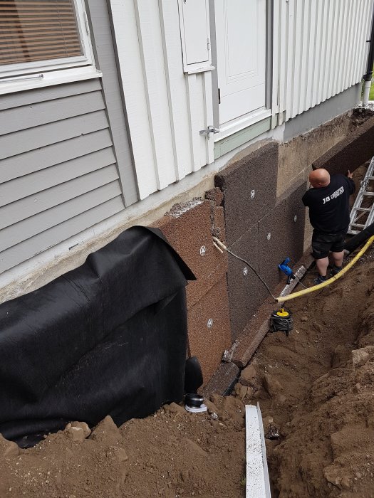 Person gräver för husdränering vid källarvägg med ny isolering och gammalt tegelrör.