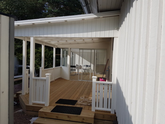 Nybyggd altan med tak intill en vit husfasad, trappsteg ner och utemöbler synliga.
