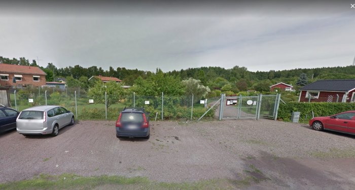Bild på en parkeringsplats med två bilar, en häck och grindstolpar i förgrunden och röda hus i bakgrunden.