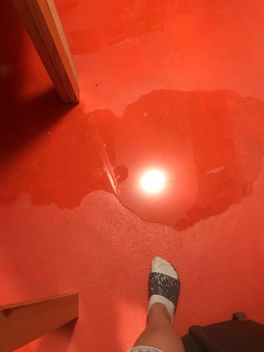 Vattenpöl på ett rött golv med en fot i förgrunden.