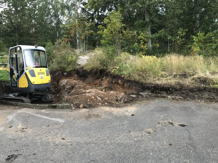 Minigrävare vid uppriven slänt med stor sten synlig i jordhögen för utbyggnad av parkeringsyta.