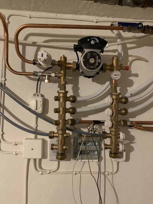 Golvvärmesystem med kopparledningar, en cirkulationspump och olika justeringsventiler på en vit vägg.
