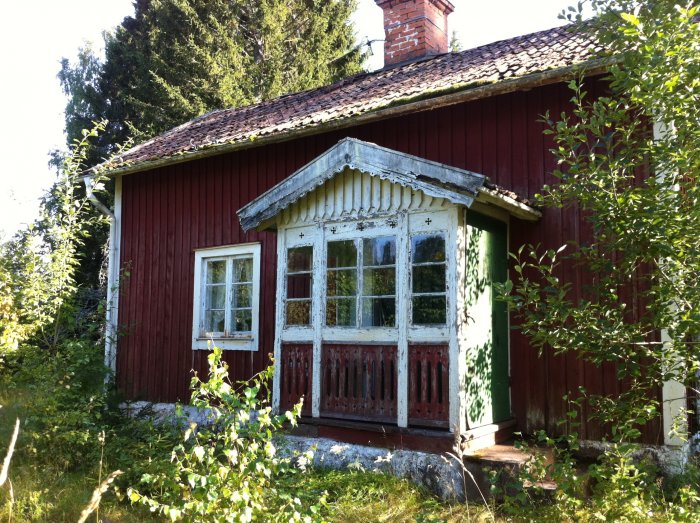 Gammalt rött torp med vitmålad veranda och slitna fönster, beläget i en grönskande omgivning.