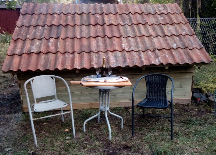 Nybyggd latrinkomposteringsbyggnad med rött tak och festdukning med proseccoflaska och två glas.