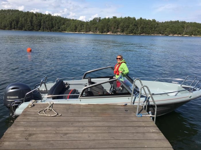 Person i grönt och solglasögon sitter i en Yamarin Cross 57br motorbåt förtöjd vid en träbrygga.