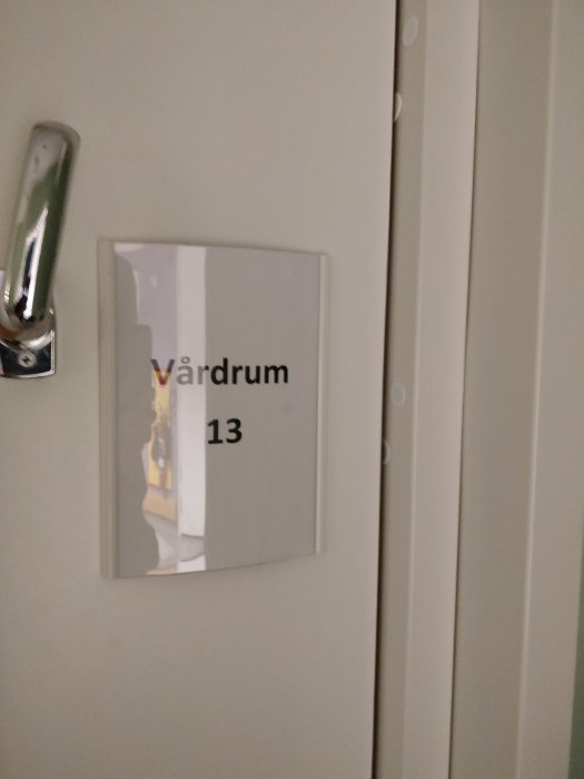 Vit dörr med skylt som säger "Vårdrum 13" på en sjukhuskorridor.