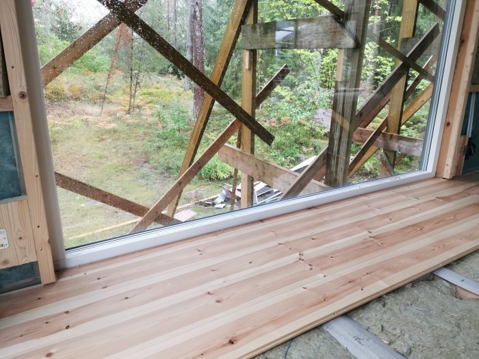 Nyinstallerade furugolvplank i en stuga med stort fönster med utsikt mot skogen.